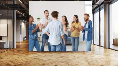 Joyful enthusiastic friends applaud man expressing their respect and congratulating him on victory. Young multiracial people clapping hands to male friend while standing in living room at home. Wall mural