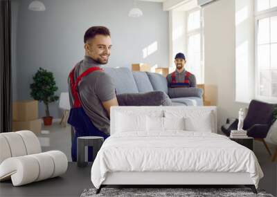 loaders at work. Male workers of furniture delivery service move sofa to customer's new apartment. Two strong men in blue and red overalls are carrying furniture. Loading service concept. Wall mural