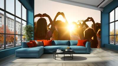 Love summer. Group of preteen children with raised hands showing heart shape in summer sun rays. Angled back view of children making heart shapes with their hands against blue sky background. Wall mural