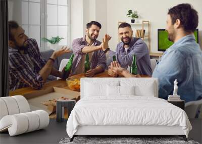 Male friends enjoying good time at fun get-together on weekend. Group of happy young men sitting at table at home, drinking beer, eating pizza, watching TV, playing games and telling funny stories Wall mural