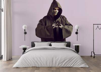 Man dressed in scary Halloween costume. Studio portrait of Mr Death with skeleton skull makeup on face wearing black cape with hood looking at camera while standing isolated on light purple background Wall mural