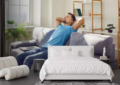 Man relaxing on a comfortable couch at home. Young guy in a blue T shirt sitting on a gray couch with his hands behind his head and a laptop beside him. Relaxation, break, pause, free time concept Wall mural