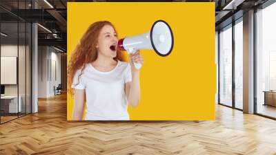 Marketing, seasonal sales and discounts. Expressive girl using megaphone to announce discounts and sales isolated on yellow background. Red-haired curly girl in white t-shirt speaks into loudspeaker. Wall mural