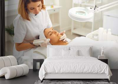 Microdermabrasion skincare in beauty salon. Smiling woman dermatologist making skincare procedure of ultrasound Microdermabrasion with special metal machine for relaxing young woman Wall mural