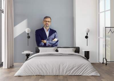 Middle aged businessman sitting at wall with folded hands. Portrait of attractive man wearing glasses and formal wear posing in office. Successful leader, executive director concept Wall mural