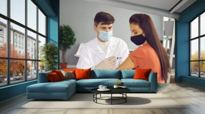 Middle aged confident man doctor in white uniform and medical mask making injection from coronavirus for young woman patient in medical clinic. Vaccination and vaccine agaist covid-19 concept Wall mural