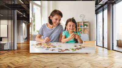 Mom and little daughter put together puzzles while sitting at a table in the room. Wall mural