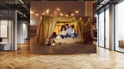 Mother with two daughters reading book and telling story hiding in tent. Happy family having fun together before going to bed in dark evening. Home living room with flashlight decoration Wall mural