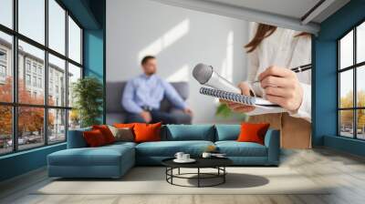 Newspaper journalist or TV correspondent gets ready for interview. Young woman with notebook, pen and microphone in hands standing in studio with famous guest in background. Press, television concept Wall mural