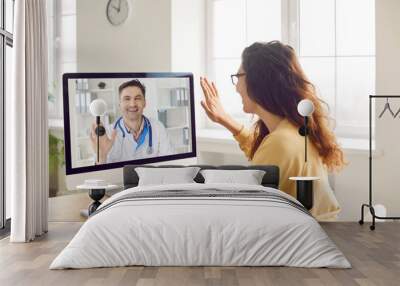 Patient says hello to online doctor when they start medical consultation via video call. Woman sitting in front of computer screen and waving her hand to greet her remote doctor. Telemedicine concept Wall mural