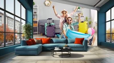 Playful funny couple having fun on air mattress at home, imagining themselves on summer vacation. Man and woman in summer hats and sunglasses are having fun at home among colorful suitcases. Wall mural