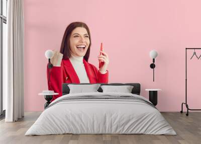 Portrait happy joyful excited beautiful business woman in red suit on solid pastel pink empty blank copy space studio background, holding phone, celebrating winning good deal and shouting Yes, Hooray Wall mural