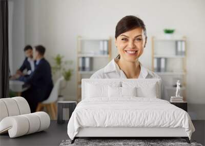 Portrait of a happy smiling confident young business woman wearing white shirt looking at camera with a company employees talking and working at desk in background in modern office. Wall mural