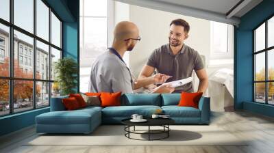 Portrait of a happy smiling young man patient in medical office listening to doctor holding report file with appointment and giving consultation during medical examination in clinic. Wall mural