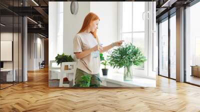 Portrait of a happy smiling young redhead woman wearing casual clothes spraying water on a green plant standing in living room at home. Gardening and caring for houseplants concept. Wall mural