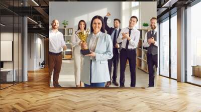 Portrait of a happy smiling young woman winner holding the trophy award for success in business standing in office with a team of people colleagues applauding for company best employee reward. Wall mural