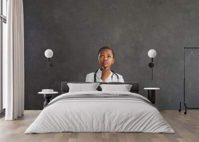 Portrait of a serious dark-skinned female doctor with a pensive expression on a gray background. Pensive woman in a medical gown looks at the free space for the text above her head. Banner. Wall mural