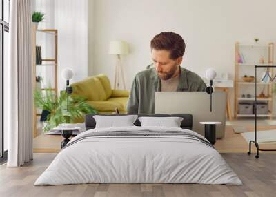 Portrait of a young business man in casual clothes working on a laptop computer at office or at home and writing at the desk making notes or calculations. Remote and freelance work concept. Wall mural