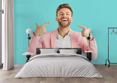 Portrait of a young handsome bearded man wearing bright pink suit and white shirt pointing at himself with index finger smiling positive and cheerful at camera isolated on studio blue background. Wall mural