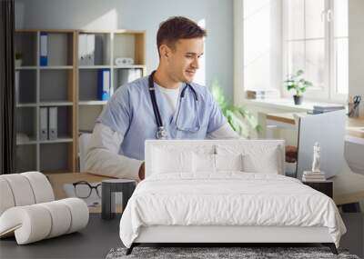 Portrait of a young male doctor or nurse in blue medical uniform sitting at the desk and working in office. Man physician wearing stethoscope in clinic using laptop typing and writing in notepad. Wall mural