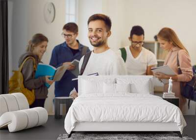 Portrait of cheerful male university student in classroom. Handsome young man with happy face expression holding textbook and smiling at camera while his classmates are reading books in background Wall mural