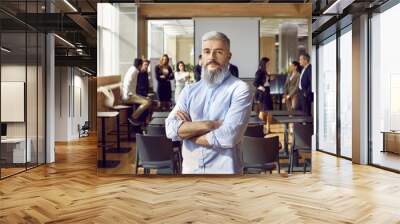 Portrait of experienced team coach and business professional. Confident man with white hair and gray beard standing with arms folded in office after corporate training meeting with group of employees Wall mural