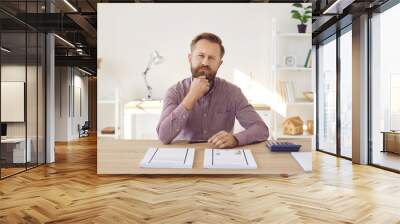 Portrait of focused pensive businessman looking at camera while sitting in office. Man sits at table with documents and calculator and thinks about plan to increase company's profits. Wall mural