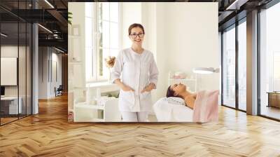 Portrait of friendly female woman beautician, aesthetic nurse or masseuse at her workplace. Smiling female beauty salon worker standing next to her client lying on couch in bright office. Wall mural