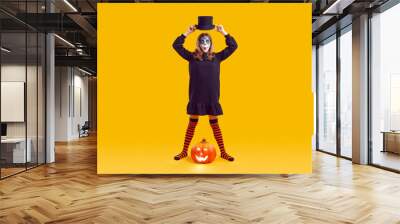 Portrait of funny teenage girl with Halloween makeup looking surprised on orange background in studio. Child in black dress and striped leggings holds hat over his head while standing next to pumpkin. Wall mural