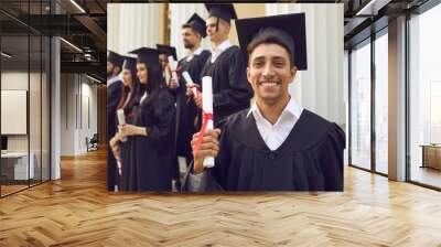 Portrait of happy confident graduate guy dressed in black mantle with diploma in hands on university stairs. Student looks at the camera and smiles sincerely. Wall mural