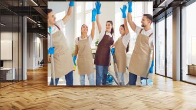 Portrait of happy excited smiling young team of janitors in uniform from professional cleaning service standing in a circle with hands up finishing work in clients apartment. Housework concept. Wall mural