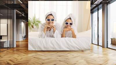 Portrait of happy good looking mother and pretty little daughter wearing trendy sunglasses and head turban towels lying on bed and smiling at camera while enjoying beauty facial skincare on spa day Wall mural