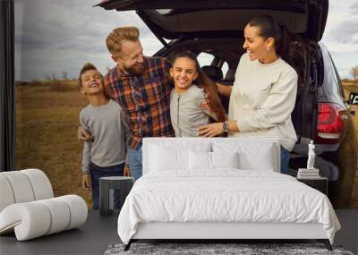 Portrait of happy smiling family of four with two kids boy and girl having fun and hugging standing near the trunk of their car enjoying weekend in nature. Vacation and road trip concept Wall mural