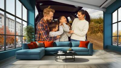 Portrait of happy smiling family of four with two kids boy and girl having fun and hugging standing near the trunk of their car enjoying weekend in nature. Vacation and road trip concept Wall mural