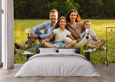 Portrait of happy smiling family with son and daughter sitting on green grass in the summer park and looking cheerful at camera. Mother, father with two kids enjoying sunny day together. Wall mural