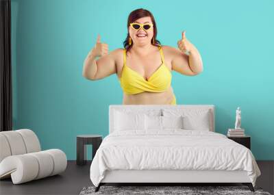 Portrait of happy smiling fat woman wearing yellow swimsuit showing thumb up sign isolated on a blue studio background. Travelling, body positive, vacation and summer journey concept. Wall mural