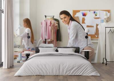 Portrait of happy young female seamstress or tailor at work. Cheerful beautiful Caucasian woman who earns living as dressmaker leaning on table in her modern studio, looking at camera and smiling Wall mural