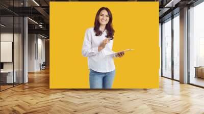 Portrait of happy young female secretary, office manager or business assistant with clipboard. Pretty woman in white shirt standing isolated on yellow background, holding clipboard and pen and smiling Wall mural