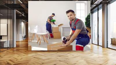 Portrait of happy young man in uniform looking at camera while packing and sealing box using sticky adhesive duct tape gun roller dispenser. Moving house, relocation company, delivery service concept Wall mural