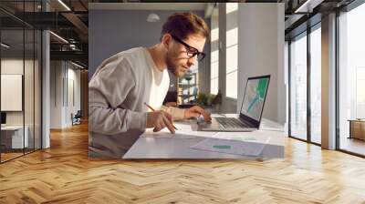 Portrait of professional cartographer looking at laptop monitor screen, working with printed cadastral map at table on workplace. Young man analyzing cadastral map searching for building plot. Wall mural