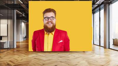 Portrait of redhead bearded chubby smiling man in glasses and funky outfit yellow shirt and red jacket looking aside by eyes on copy space on yellow background. Banner for advertisement, marketing. Wall mural