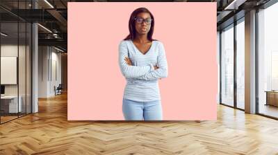 Portrait of serious female college or high school student. Young African American woman or teenage girl wearing blue long sleeve top, jeans and glasses standing, arms folded, on pastel pink background Wall mural