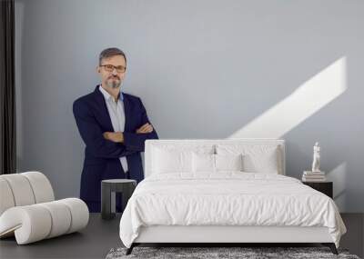 Portrait of successful middle-aged business man standing in confident pose against gray wall. Caucasian man in stylish business suit stands with his hands folded. Concept of successful business people Wall mural