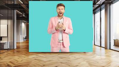 Portrait of surprised excited man in modern stylish pink suit holding mobile phone on a studio blue background. Young amazed guy with a shocked face expression looking at the camera. Wall mural