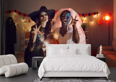 Portrait of two beautiful young women dressed up in spooky Wicked Witch and Dead Bride costumes with ugly makeup on faces doing claw hand gesture, hissing, making grimaces, wishing you Happy Halloween Wall mural