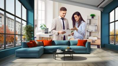 Portrait of two smiling business people man and woman standing in office at their workplace and looking through financial documents. Employees discussing work project or company finances. Wall mural