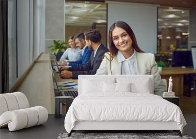 Portrait of young happy smiling business woman sitting in modern open plan office at same desk with coworkers with laptop and looking cheerful at camera. Female company employee working on workplace. Wall mural