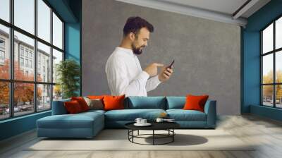 Profile view portrait of happy satisfied young man using mobile phone standing isolated on grey copyspace background. Cheerful guy gets good news. Boyfriend receives love message from girlfriend Wall mural