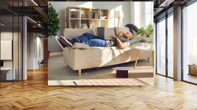 Relaxed man enjoying his day off at home. Happy guy watching a good movie on his laptop while resting on the sofa in the living room interior, with a book on the floor and a snack on the coffee table Wall mural