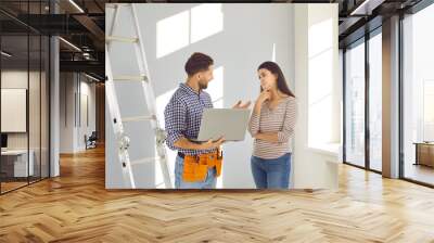 Repairman with modern laptop talking to young homeowner girl. Male worker with tool belt standing by step ladder, holding notebook computer and explaining to woman what he has to repair in the house Wall mural